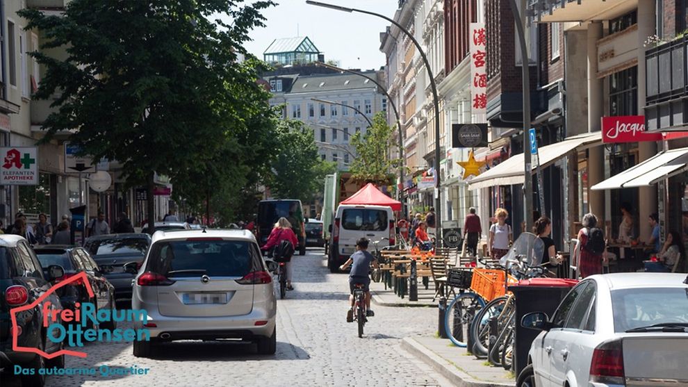 Die Ottensener Hauptstraße mit Blick Richtung Reitbahn. Rechts und links neben der Fahrbahn parken Autos. Auf der Straße fährt ein silberner Kombi. Daneben ein Junge auf dem Fahrrad mit Fahrradhelm.