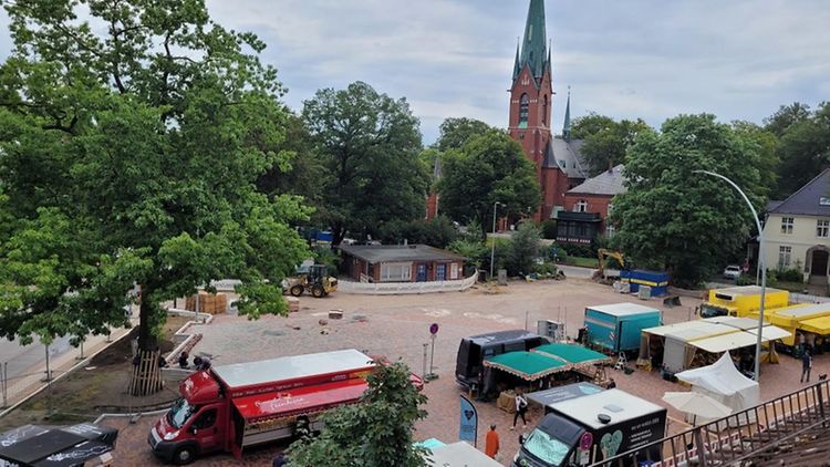  Der Blankeneser Marktplatz mit Blick in Richtung Marktkirche. Auf dem Platz stehen vereinzelt Marktstände. Die großen Bäume um und auf dem Platz tragen dichtes, grünes Laub.