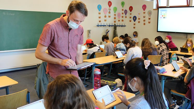  Ein Lehrer steht im Klassenraum und tippt etwas auf seinem Tablet, vor ihm sitzen Schülerinnen und tippen ebenfalls auf ihren Tablets, sie haben Masken auf. 