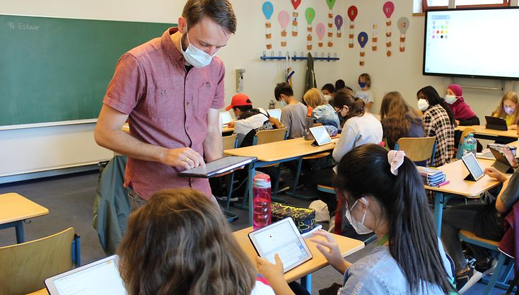 Ein Lehrer steht im Klassenraum und tippt etwas auf seinem Tablet, vor ihm sitzen Schülerinnen und tippen ebenfalls auf ihren Tablets, sie haben Masken auf. 