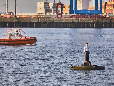  Vor Containern im Hafen steht ein Bojenmann von Stephan Balkenhol auf dem Wasser