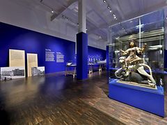  Ein Ausstellungsraum der Ausstellung erste Dinge mit blauen Wänden und einer göttlichen Figur auf einem Löwen in einer Vitrine.