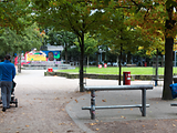 Ein Mann schiebt einen Kinderwagen durch einen begrünten Park, indem sich Sitzmöglichkeiten und Bänke befinden. 