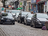  Eine Einbahnstraße mit Kopfsteinpflaster, auf der sich links und rechts parkende Autos befinden sowie ein fahrendes Auto.