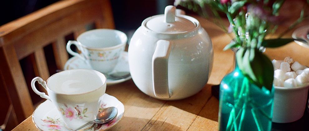  Zwei filigrane Teetassen und eine Teekanne aus weißem Porzellan auf einem Holztisch. Daneben stehen pinke Blumen in einer dunkelgrünen Vase und ein weißes Schälchen mit Zucker.