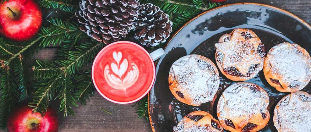  Ein Heißgetränk mit rotem Milchschaum und runde Weihnachtstörtchen mit Puderzucker auf einem Teller. Links im Bild Tannenzweige, Kiefernzapfen und Äpfel als Deko.