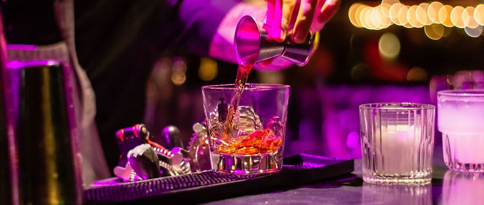  Eine Hand, die Likör in ein Whiskyglas mit Eiswürfeln kippt. Der Hintergrund ist pink beleuchtet. 