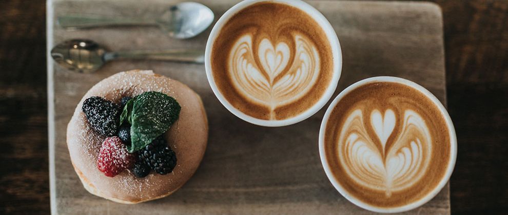  Ein Gebäck mit Zucker, Waldbeeren und Minzblatt dekoriert und zwei Kaffee mit Latte Art auf einem Holzbrett.