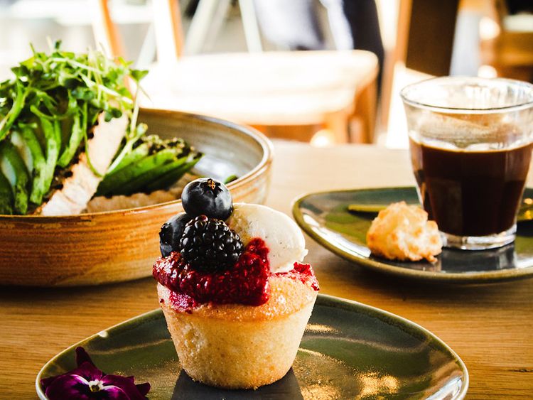  Ein Getränk und zwei Gerichte auf Keramikgeschirr. Links ein Avokadobrot, in der Mitte ein Beeren Cupcake und rechts ein Glas mit schwarzem Kaffee.