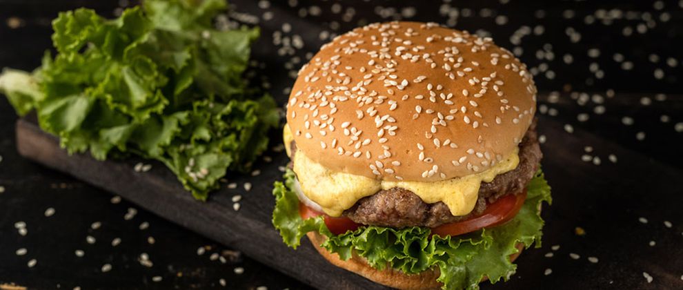  Ein Burger mit hellem Brötchen und Sesam belegt mit Salat, Tomate, Fleisch-Patty und Käse auf schwarzem Untergrund. 
