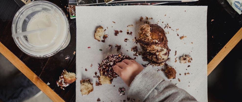  Eine Kinderhand, die einen Donut zerpflückt. Daneben steht ein Plastikbecher mit milchiger Flüssigkeit. 