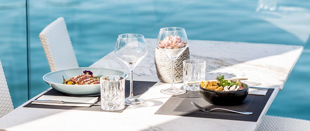  Asiatisches Essen und Thunfisch auf einem gedeckten Tisch. Im Hintergrund ist eine Glasscheibe und Wasser zu sehen..