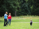  Zwei Erwachsene stehen in einem Park und warten mit offenen Armen auf ihr Kind, das ihnen entgegenläuft. 