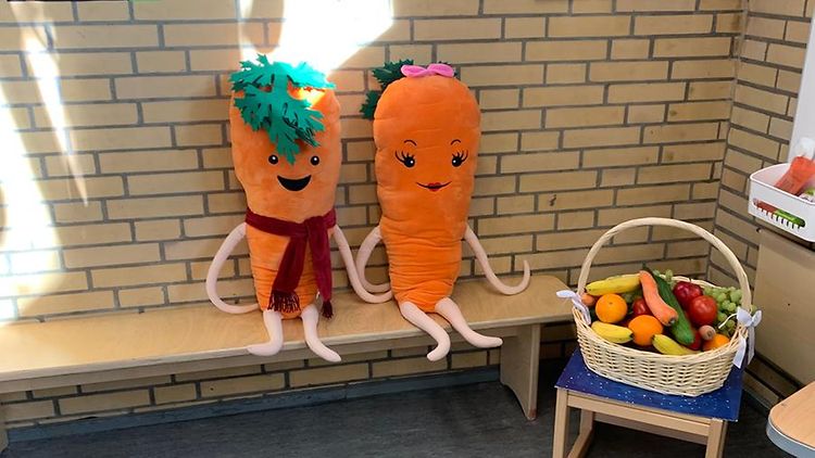  Zwei Karotten aus Stoff mit Gesichtern sitzen auf einer Bank, daneben steht ein Obstkorb.