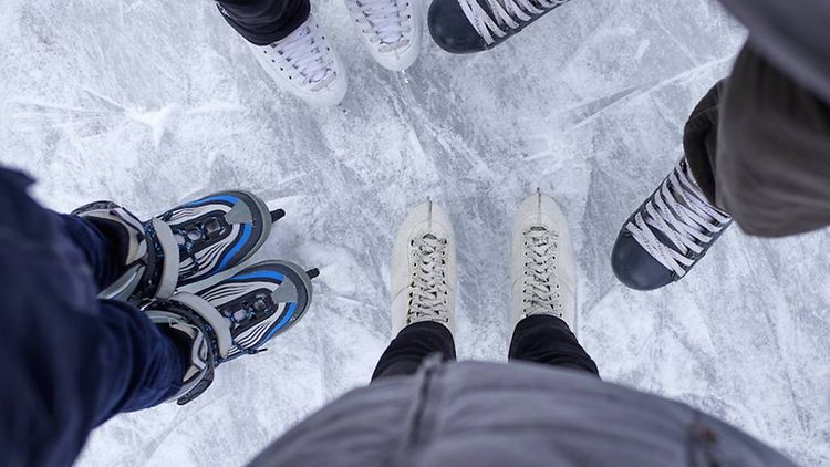  Mehrere Personen stehen mit Schlittschuhen auf dem Eis. Es sind nur die Beine zu sehen.