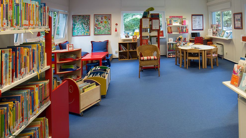 Ein Raum mit blauem Teppichboden und bunter, kindgerechter Einrichtung wie Regalen mit bunten Büchern, einer Sitzecke, einem runden Tisch mit Stühlen und selbst gemalten Bildern an den Wänden. 