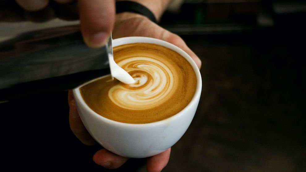 Eine Hand hält eine Kaffeetasse mit Kaffee, eine andere gießt Milchschaum aus einem silbernen Kännchen in die Tasse, sodass Latte Art entsteht.