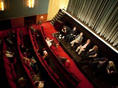  Meschen sitzen in einer Reihe auf Stühlen auf einer Bühne in einem Kinosaal und BesucherInnen sitzen in roten Kinosesseln vor der Bühne. Das Bild wurde aus der Vogelperspektive fotografiert. 