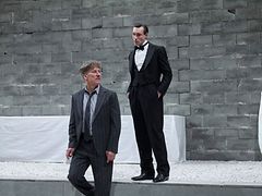  Zwei Männer in Anzügen stehen vor einer grauen Wand.