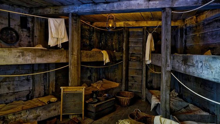 Es ist der Nachbau einer Kabine zu sehen. In dem Raum aus Holz stehen Etagenbetten und Wäscheleinen.
