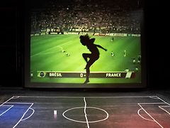  Eine Frau springt auf einem aufgemalten Fußballfeld, im Hintergrund steht ein Monitor auf dem ein Fußballspiel gezeigt wird.