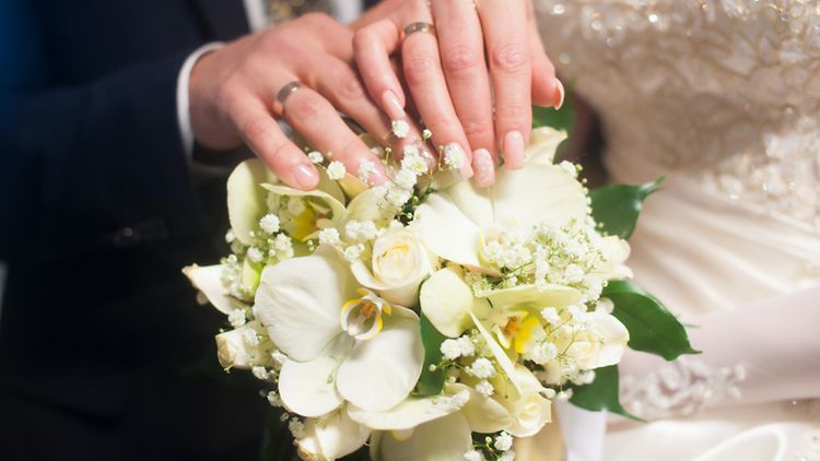  Ein Brautpaar mit Eheringen an den Händen hält einen Blumenstrauß.