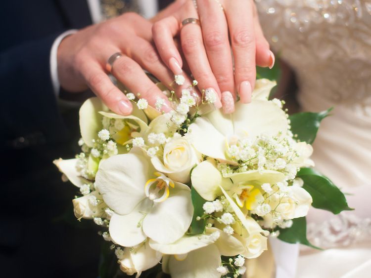  Ein Brautpaar mit Eheringen an den Händen hält einen Blumenstrauß.