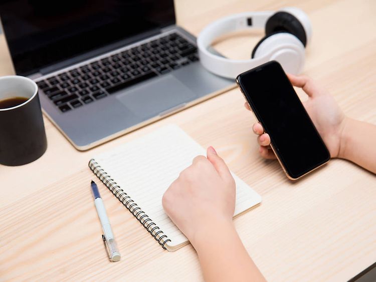  Ein Laptop, eine Kaffeetasse, Kopfhörer und ein Notizblock liegen auf einem Tisch. Hände halten ein Smartphone.