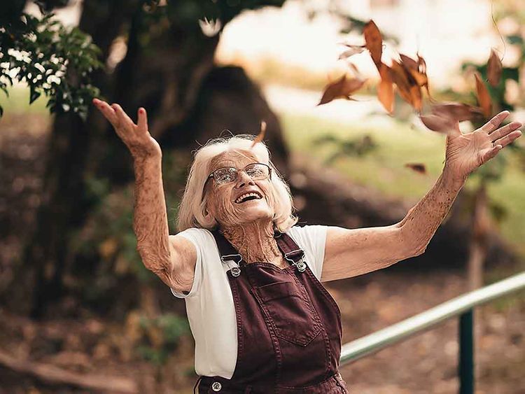  Ältere Frau steht im Garten und reißt die Arme in die Luft.