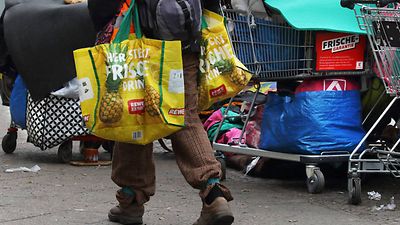  Die Beine eines Obdachlosen und im Hintergrund volle Einkaufswägen.