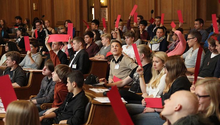 Jugendliche im Plenarsaal stimmen bei einer Sitzung von „Jugend im Parlament“ ab. Dazu halten sie rote Zettel nach oben.