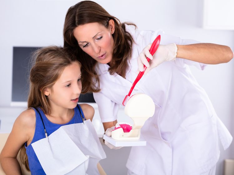  Eine Frau in Arztkleidung zeigt einem Mädchen anhand eines Modellgebisses und einer großen Zahnbürste das Putzen.