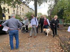  Mehrere Menschen gehen auf einem Fußweg. Ein Mann hat einen Blindenstock in der Hand, daneben steht ein Labrador.