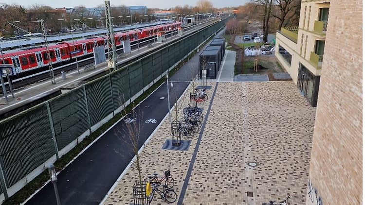  Der neu gestaltete Quartierseingangsbereich mit Eschen, mehreren Fahrrädern und einer Bahn am linken Bildrand.
