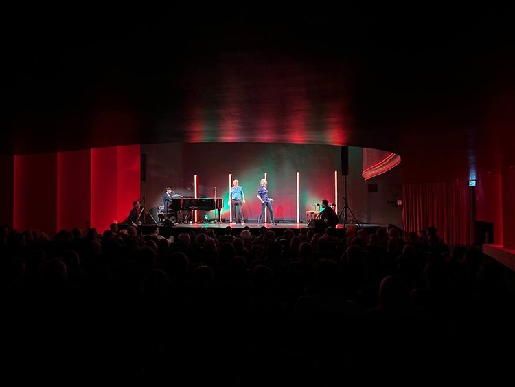  In einem dunklen Raum stehen zwei Menschen vor einem Publikum auf einer Bühne.