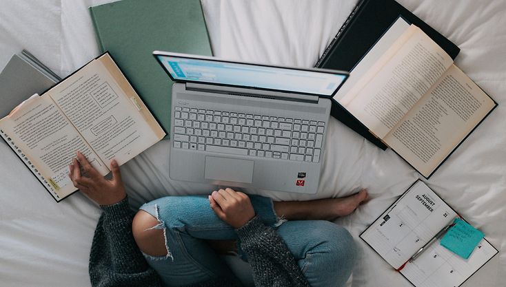 Woman sitting on a bed in front of her laptop and surrounded by different books and notebooks