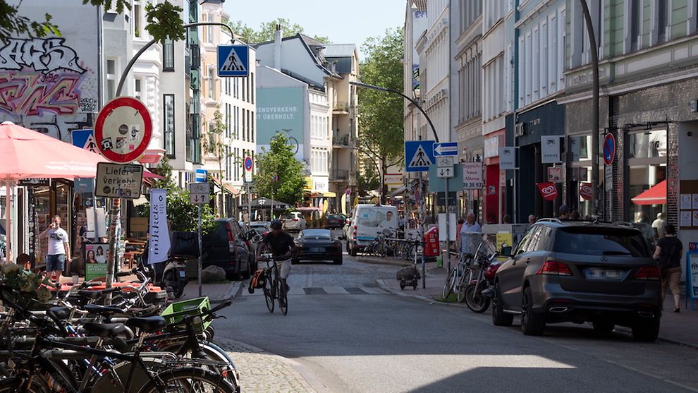 Straße in Ottensen voller Verkehrsteilnehmer, parkenden Fahrrädern und Autos am Straßenrand sowie Passanten.