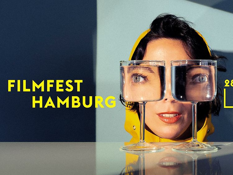  Plakat für das Filmfest Hamburg 2023. Eine Frau, die durch zwei mit Wasser gefüllte Gläser schaut.