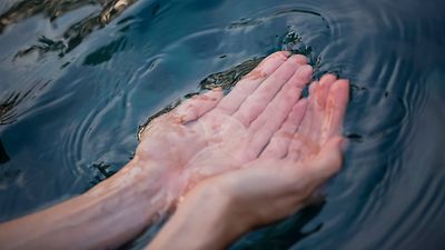  Im Zentrum des Bildes sind zwei Hände mit aneinander gehaltenen, geöffneten Handflächen zu sehen. Die Hände sind in eine dunkelblaue Wasserquelle mit leichten Wellenbewegungen getaucht. 
