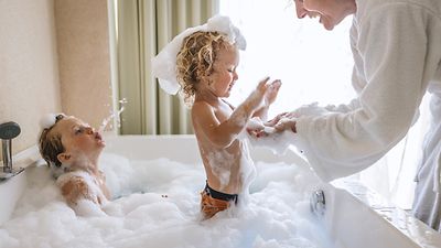  Zwei kleine Kinder spielen in einer weißen, mit Schaum gefüllten, Badewanne. Das Kind auf der linken Seite sitzt im Schaumbad und spuckt Wasser in einem Strahl in die Luft.