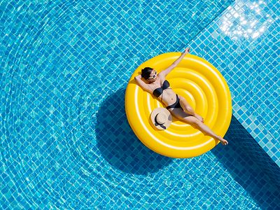  Ein Pool mit unterschiedlichen blauen Kacheln aus der Vogelperspektive: Auf der rechten Seite liegt eine Frau in einem schwarzen Bikini und mit einer Sonnenbrille auf einer runden, gelben Luftmatratze