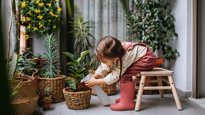  Ein kleines Mädchen in rosa Latzhose und Gummistiefeln sitzt vornübergebeugt auf einem Holzhocker und gießt eine Pflanze mit einer Sprayflasche. Im Hintergrund stehen mehrere Pflanzen in Basttöpfen.