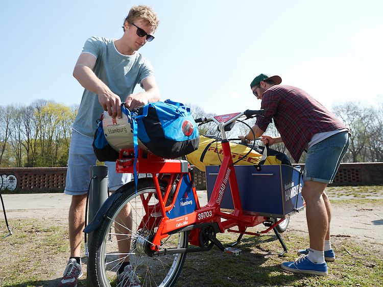  Zwei Männer bepacken ein rotes Lastenrad.