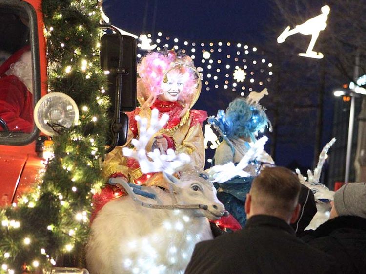  Verkleidete Figuren laufen auf der Weihnachtsparade mit und stehen auf geschmückten Fahrzeugen. An den Straßenseiten stehen Zuschauende. 