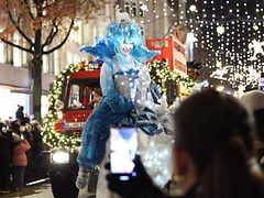  Verkleidete Figuren laufen auf der Weihnachtsparade mit und stehen auf geschmückten Fahrzeugen. An den Straßenseiten stehen Zuschauende. 