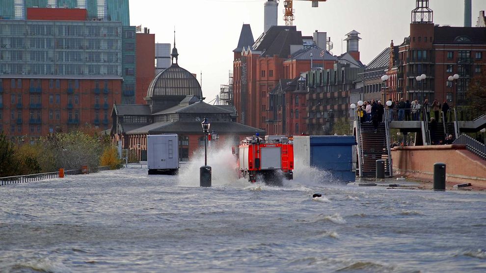  Die Elbe ist über ihre Ufer getreten und überflutet die Fläche des Fischmarktes. Ein Feuerwehrauto fährt durch das aufspritzende Wasser. Schaulustige beobachten die Szene von einer Brücke aus.