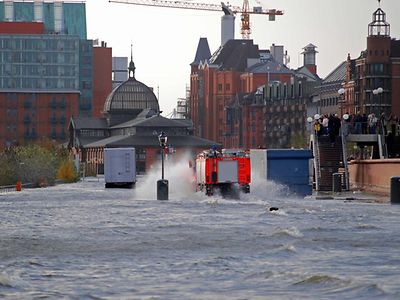  Die Elbe ist über ihre Ufer getreten und überflutet die Fläche des Fischmarktes. Ein Feuerwehrauto fährt durch das aufspritzende Wasser. Schaulustige beobachten die Szene von einer Brücke aus.