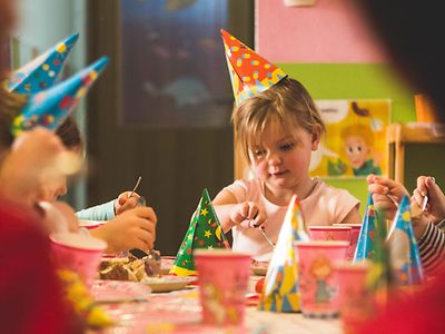 Mehrere Kinder sitzen an einem Tisch und essen Kuchen. Sie tragen spitze Papphüte mit bunten Mustern.