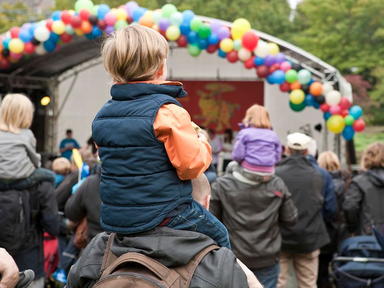  Kinder sitzen auf den Schultern von Erwachsenen und schauen auf eine Bühne, die mit bunten Luftballons behangen ist.