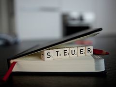  Ein ledergebundener schwarzer Kalender mit rotem Lesezeichenband liegt auf einem Tisch. Unter dem Deckel klemmen Scrabble-Würfel, auf denen das Wort "Steuer" zu lesen ist.